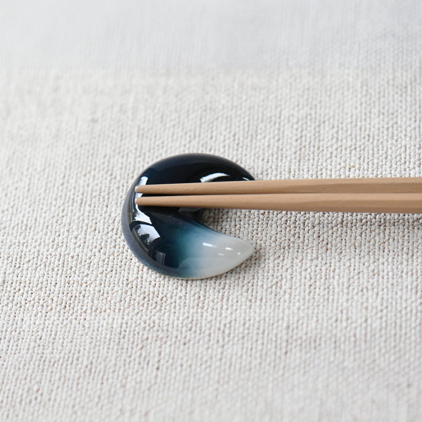 Moon-shaped ceramic chopstick rest in indigo and white by Yoshito Takenishi, holding wooden chopsticks on a textured cloth.