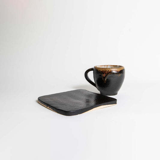 A black ceramic mug with a glossy finish sitting on a matching black ceramic plate on a white background.