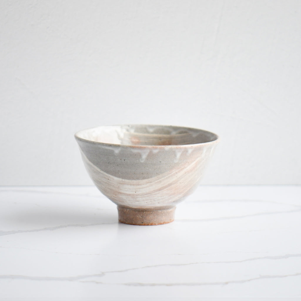  A handcrafted bowl with a gradient of gray to beige, detailed with expressive white brushstrokes and a rustic footed base, showcasing artisanal skill