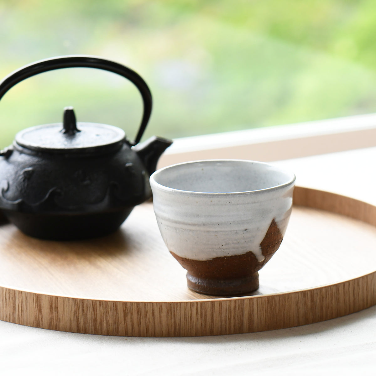 A ceramic tea cup with a white and brown glaze sits on a round wooden tray next to a black cast-iron teapot, set against a softly blurred green outdoor background.