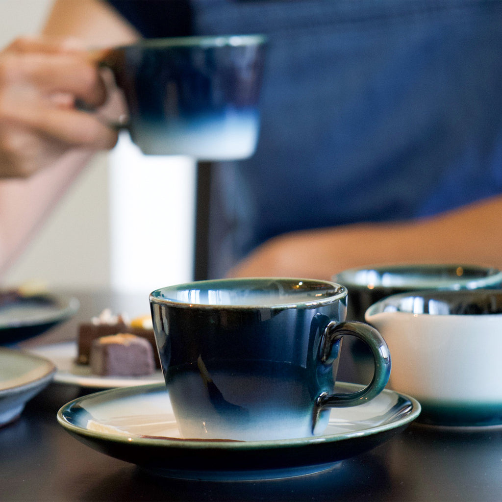 Blue coffee cup and white saucer with blue edge. A person is holding a coffee cup in the back. There are also matching creamer and sugar pots.