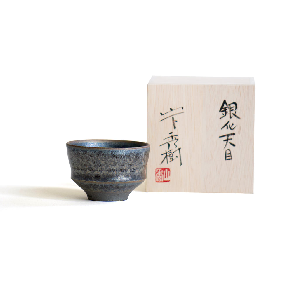 A handmade black Ginten sake bowl sitting sleekly next to a wooden box.
