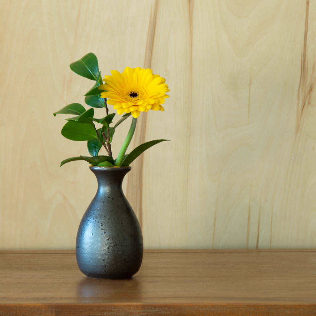 Speckled black sake container used as vase with green foliage combined with bright yellow flower